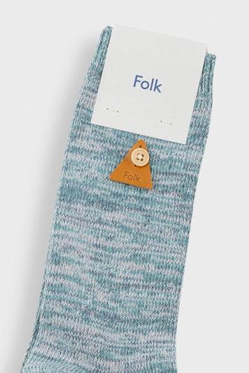 A single light blue and white heathered knit sock with a small orange-brown leather button featuring a cartoon face, shown with a white paper tag printed with the brand name 'Folk'.