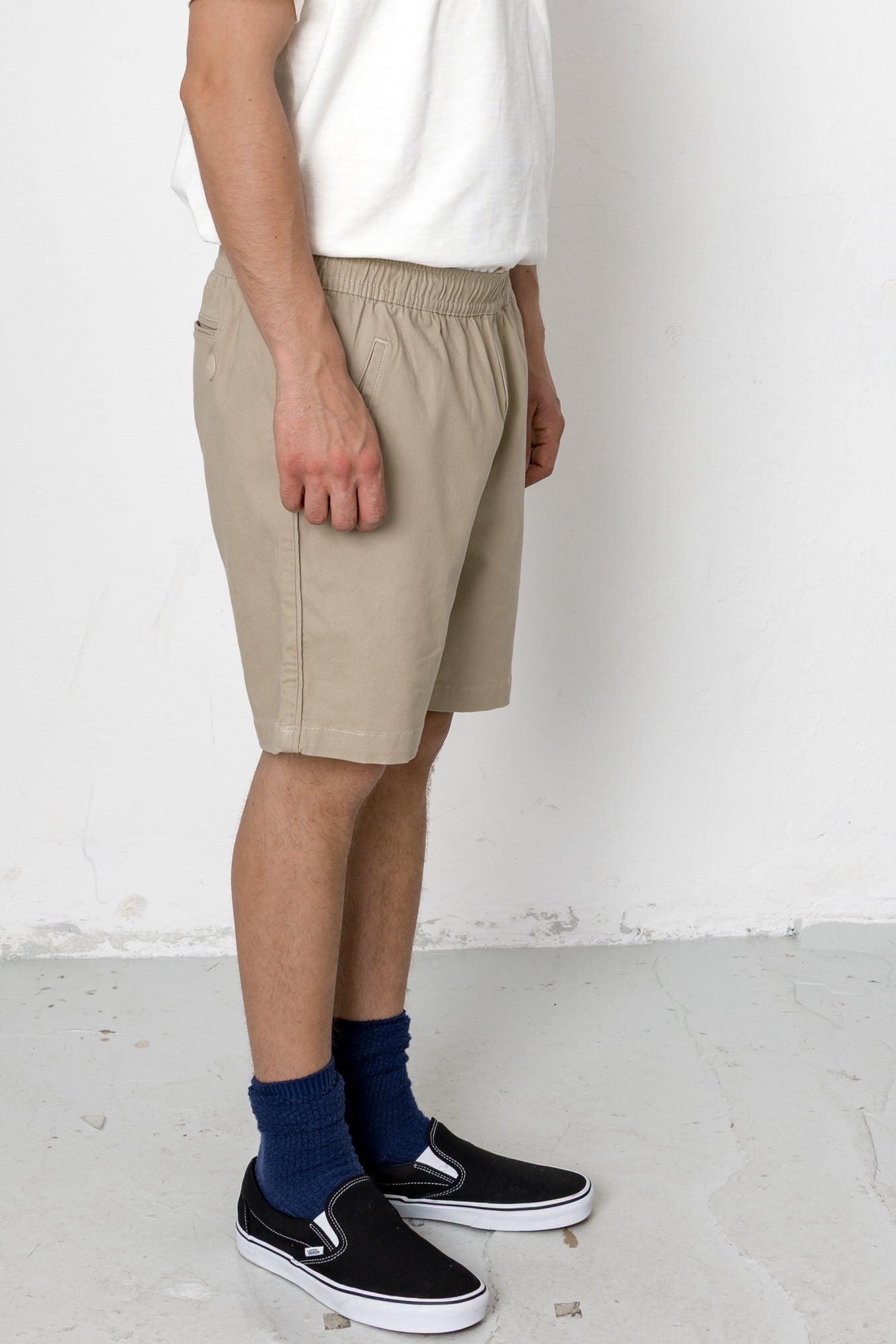 Person wearing beige shorts, white shirt, black shoes, and blue socks on a plain background