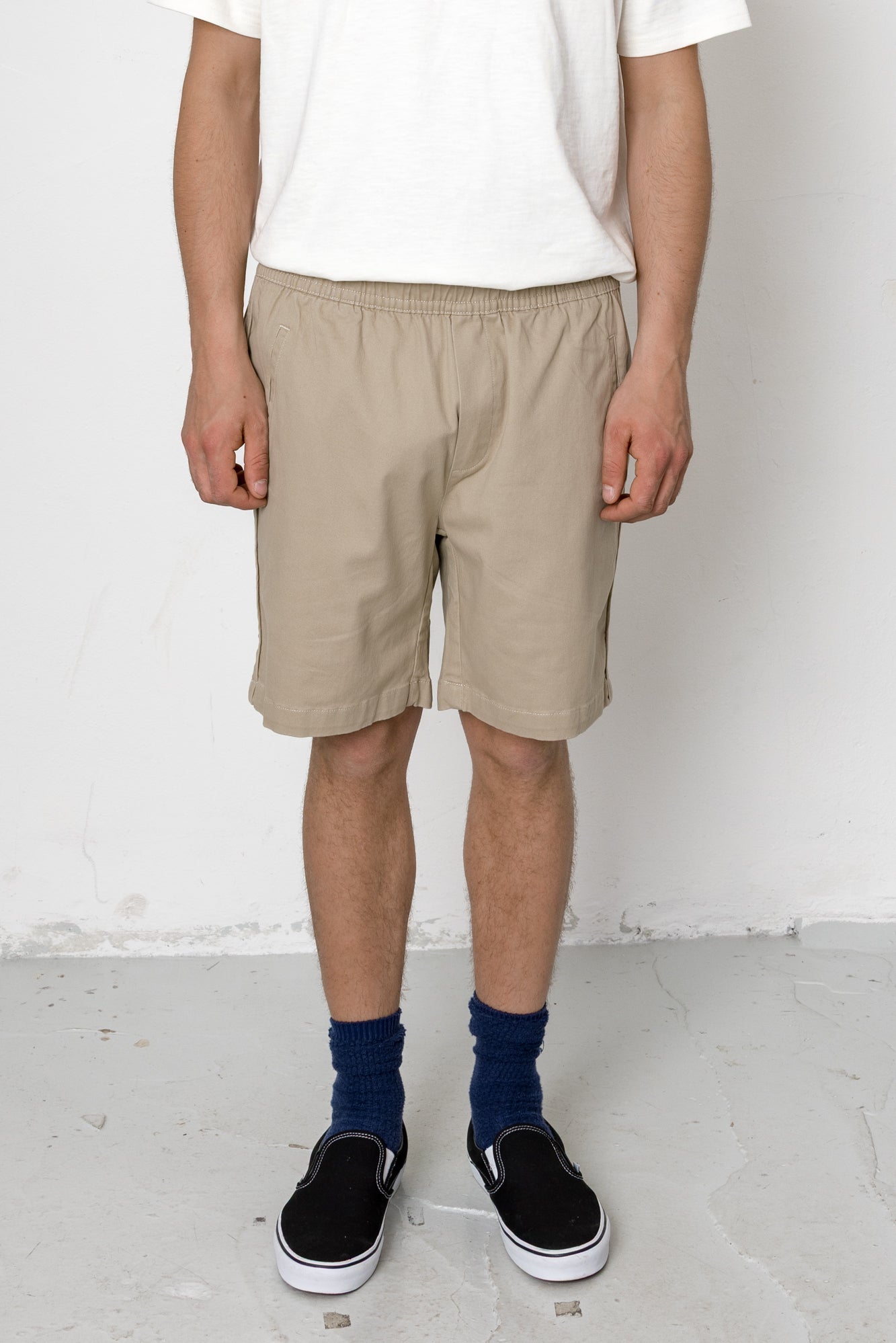 Person wearing beige shorts, white t-shirt, and black shoes on a plain background