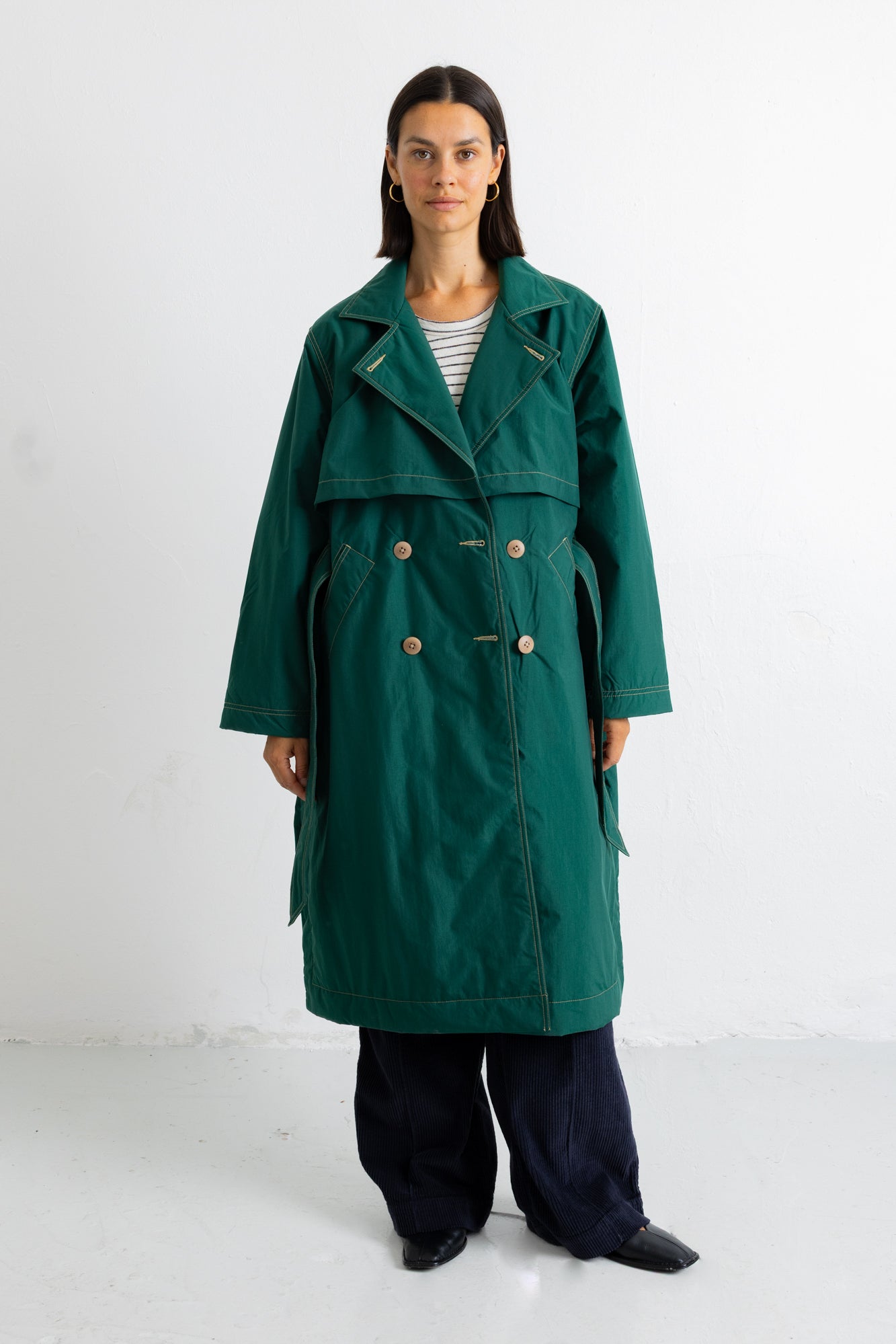 A woman stands against a plain white studio background wearing a long dark green double-breasted coat over a striped top and dark wide-leg pants.