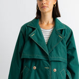 A woman wearing a dark green double-breasted utility-style coat over a white and gray striped t-shirt, photographed against a plain light gray background.