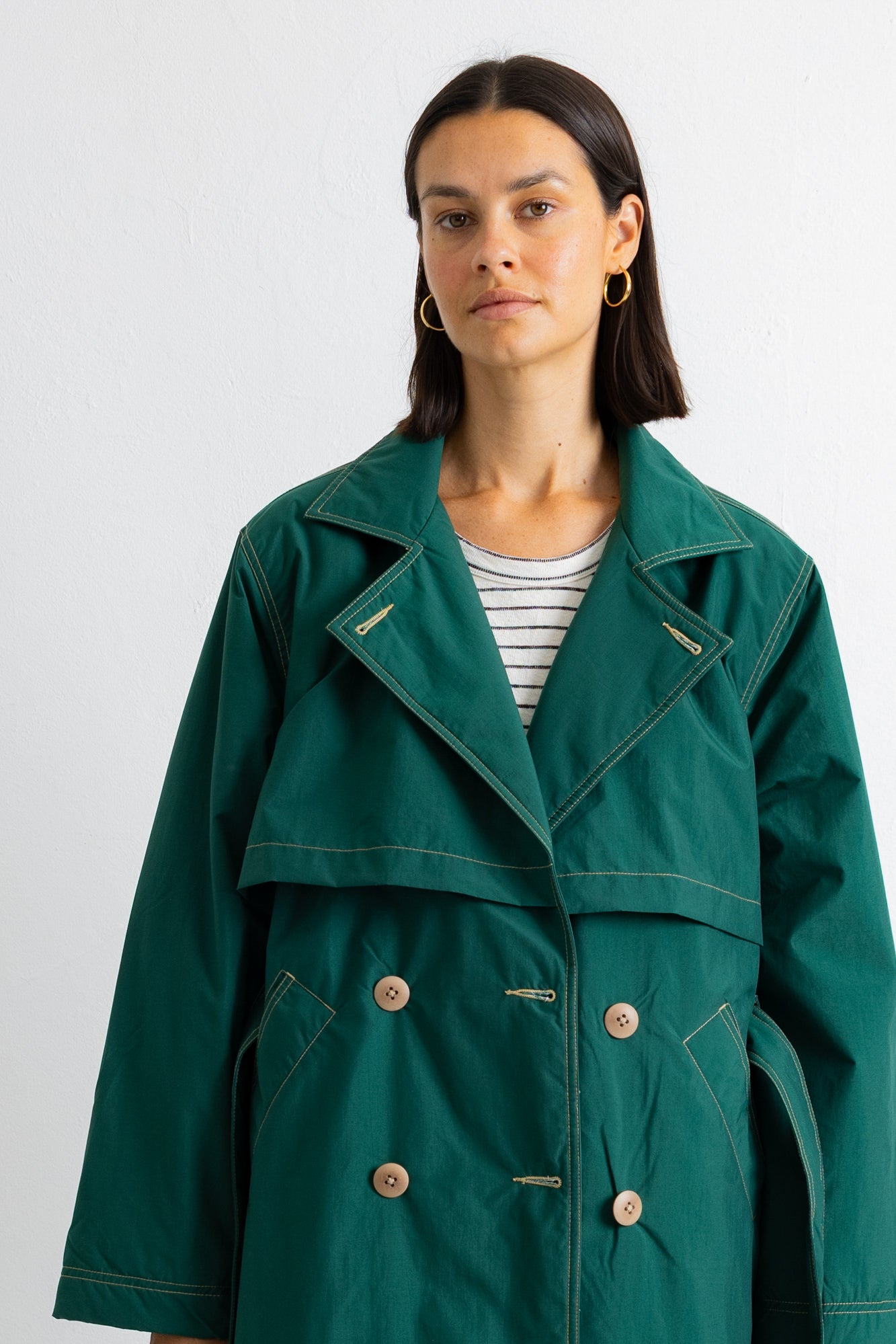 A woman wearing a dark green double-breasted utility-style coat over a white and gray striped t-shirt, photographed against a plain light gray background.