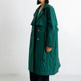 A woman modeling a long dark green trench coat with a belted waist and double-breasted buttons, paired with dark trousers and black shoes, standing against a plain white studio background.