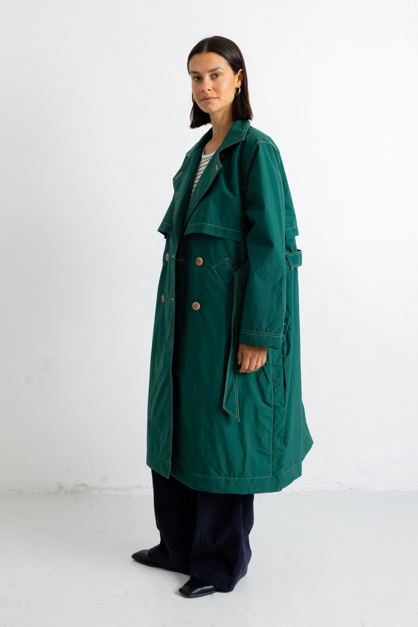 A woman modeling a long dark green trench coat with a belted waist and double-breasted buttons, paired with dark trousers and black shoes, standing against a plain white studio background.