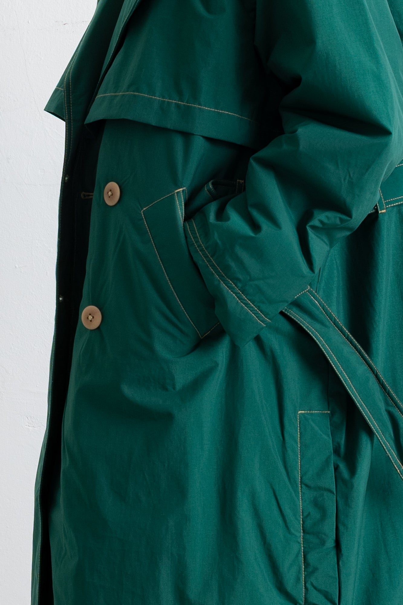 Close-up of a dark green trench coat with light tan buttons and contrast stitching, shown on a white background.