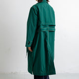 A model shown from the back wearing a long, dark green trench coat with a waist tie, paired with dark trousers, standing against a plain white studio background.