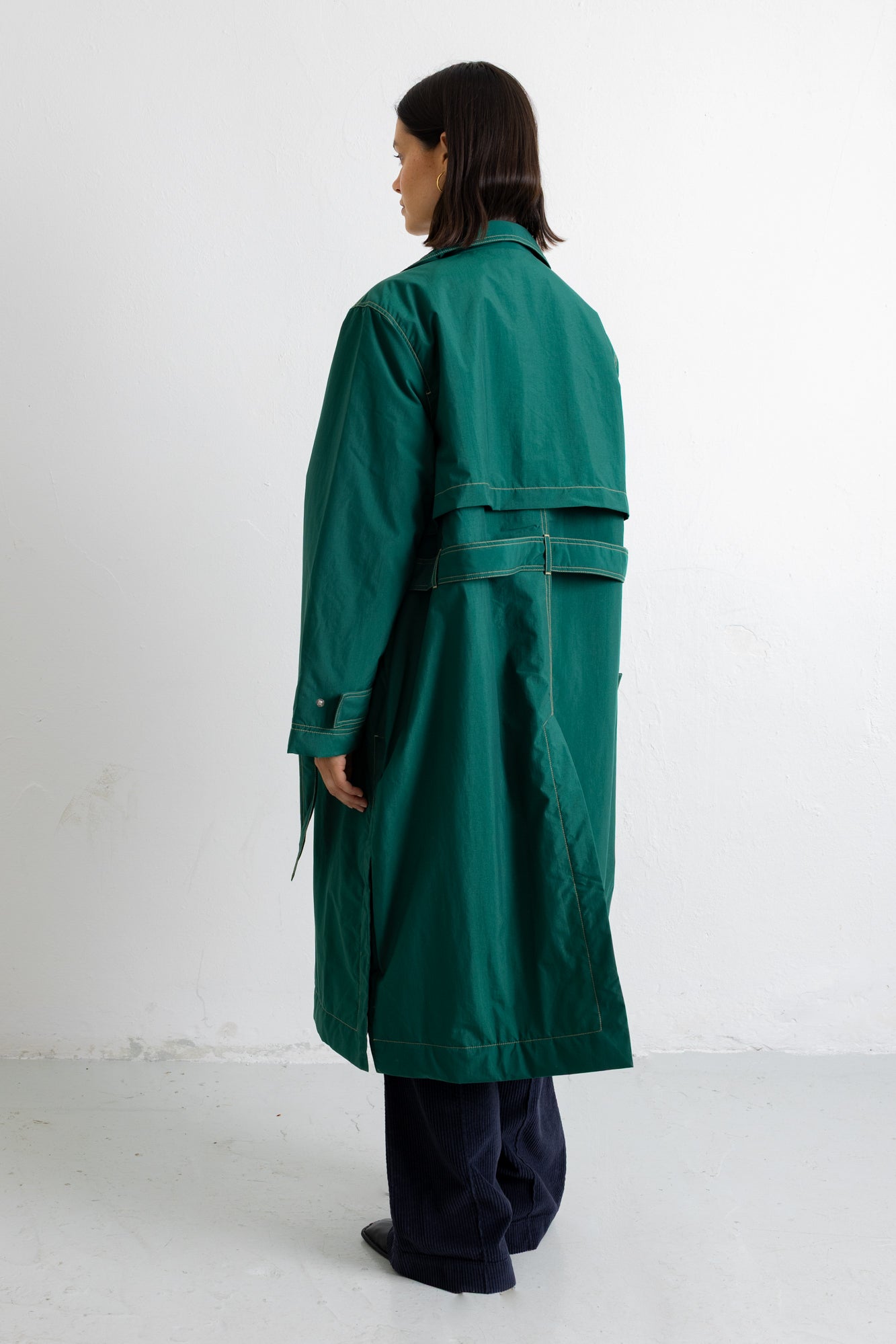 A model shown from the back wearing a long, dark green trench coat with a waist tie, paired with dark trousers, standing against a plain white studio background.