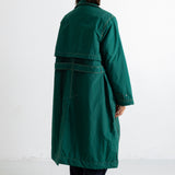 A model viewed from the back wearing a dark green mid-length coat with a wide collar and a dark belt, paired with dark trousers, against a plain white studio background.
