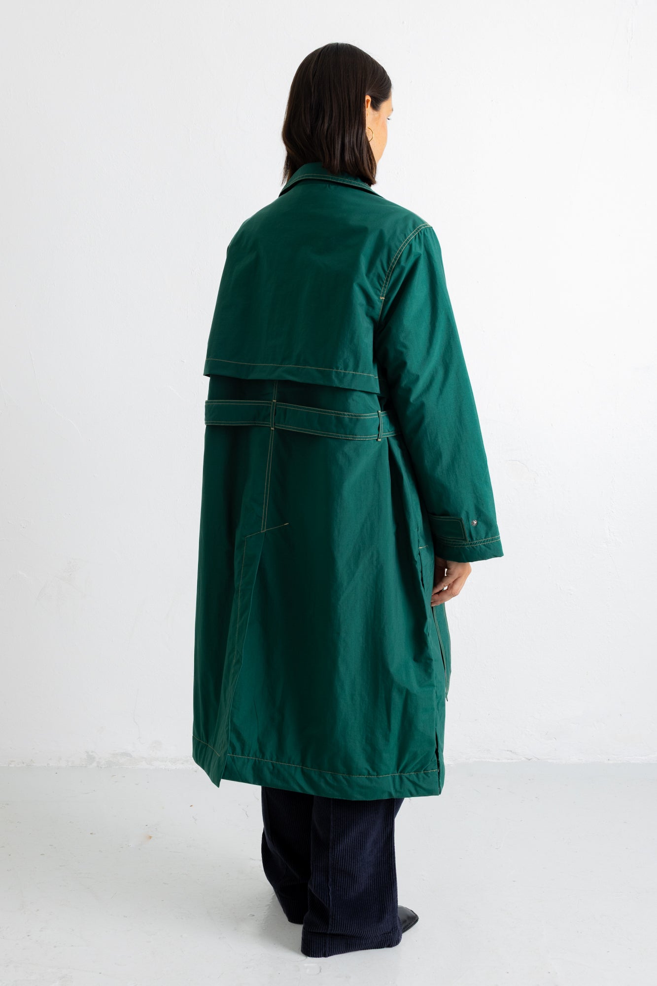 A model viewed from the back wearing a dark green mid-length coat with a wide collar and a dark belt, paired with dark trousers, against a plain white studio background.