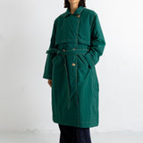 A model stands against a plain white studio background wearing a dark green belted coat over dark wide-leg trousers and black shoes.