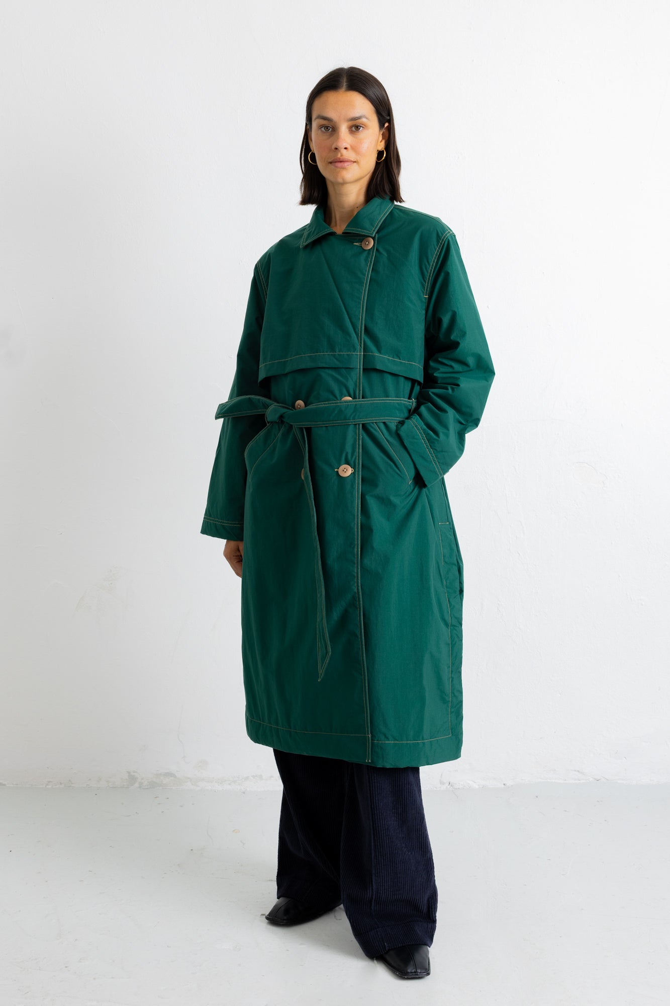 A model stands against a plain white studio background wearing a dark green belted coat over dark wide-leg trousers and black shoes.