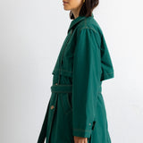 A woman shown in profile wearing a long dark green belted coat or wrap against a plain light gray studio background.