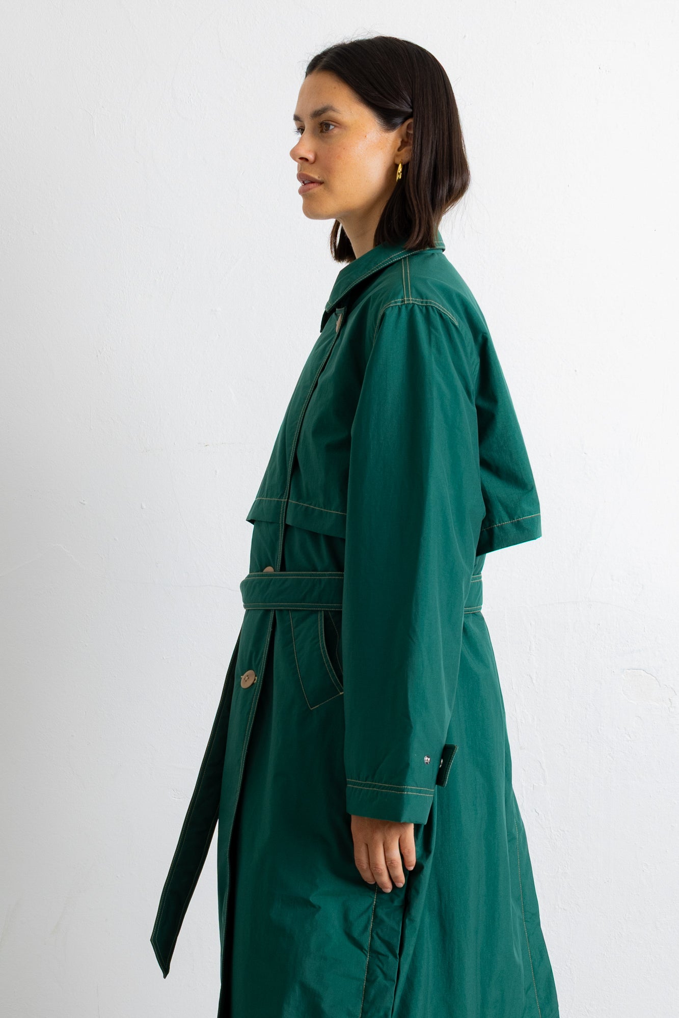 A woman shown in profile wearing a long dark green belted coat or wrap against a plain light gray studio background.