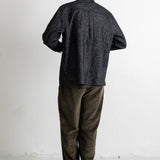 A man viewed from behind wearing a dark charcoal textured button-up shirt, olive green pants, and black canvas sneakers, standing against a plain white wall.