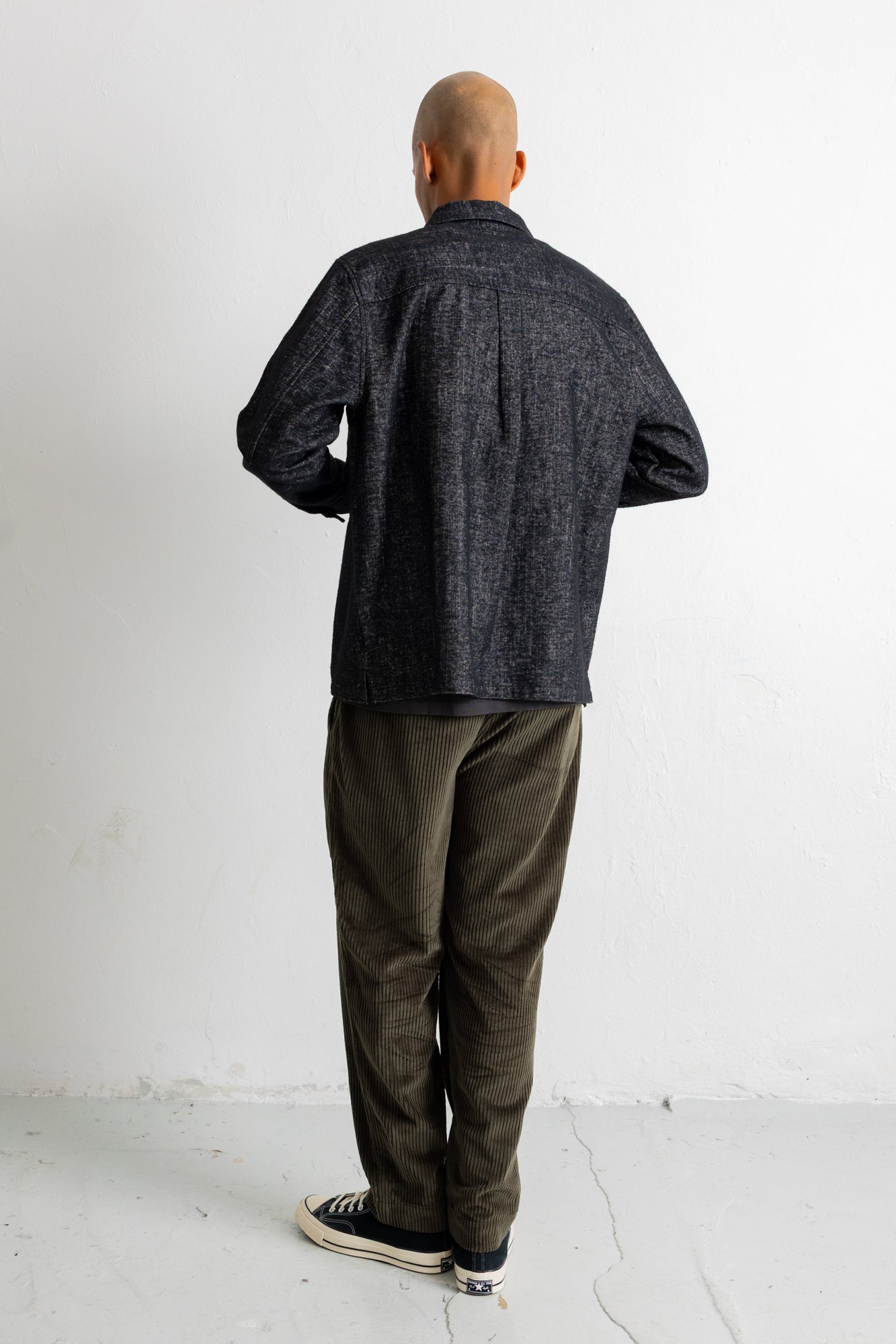 A man viewed from behind wearing a dark charcoal textured button-up shirt, olive green pants, and black canvas sneakers, standing against a plain white wall.