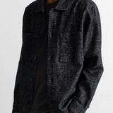 A man wearing a dark charcoal gray textured overshirt jacket over a black t-shirt and olive green corduroy trousers, photographed against a white wall.