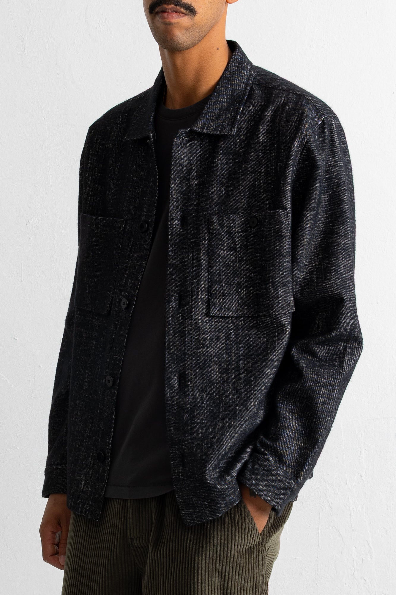 A man wearing a dark charcoal gray textured overshirt jacket over a black t-shirt and olive green corduroy trousers, photographed against a white wall.