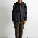 A man stands against a white wall wearing a dark charcoal textured overshirt over a black t-shirt, olive green corduroy pants, and black and white canvas sneakers.