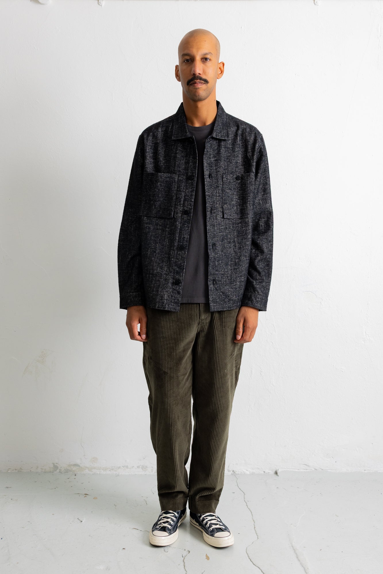 A man stands against a white wall wearing a dark charcoal textured overshirt over a black t-shirt, olive green corduroy pants, and black and white canvas sneakers.