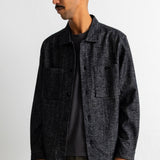 A man wearing a dark charcoal gray textured overshirt jacket over a black t-shirt and olive green corduroy trousers, standing against a plain white wall.