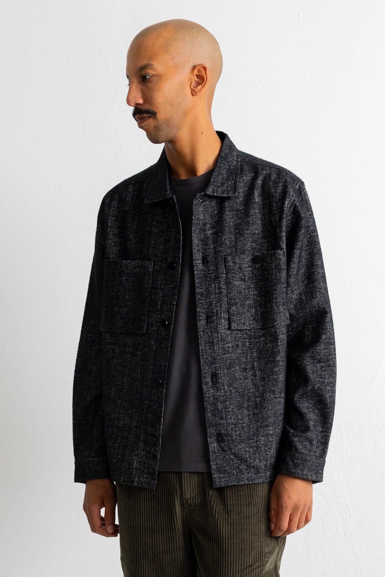 A man wearing a dark charcoal gray textured overshirt jacket over a black t-shirt and olive green corduroy trousers, standing against a plain white wall.