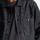 A close-up of a person wearing a dark charcoal gray textured overshirt over a black t-shirt, against a plain off-white wall.