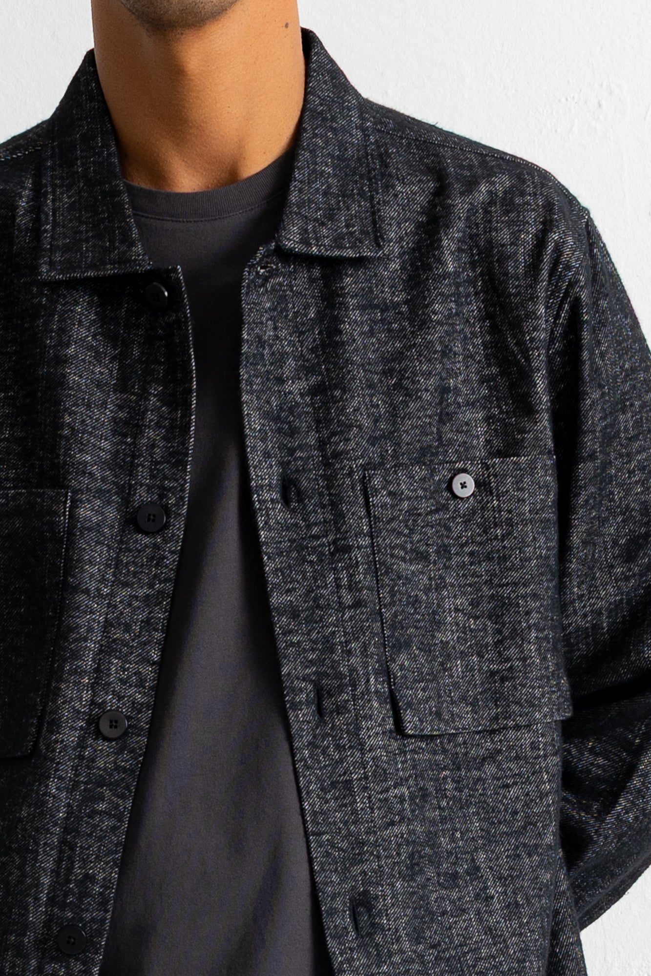 A close-up of a person wearing a dark charcoal gray textured overshirt over a black t-shirt, against a plain off-white wall.