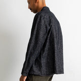 A man shown from the side profile wearing a dark charcoal gray textured long-sleeve button-up shirt and olive green corduroy trousers, standing against a plain off-white wall.