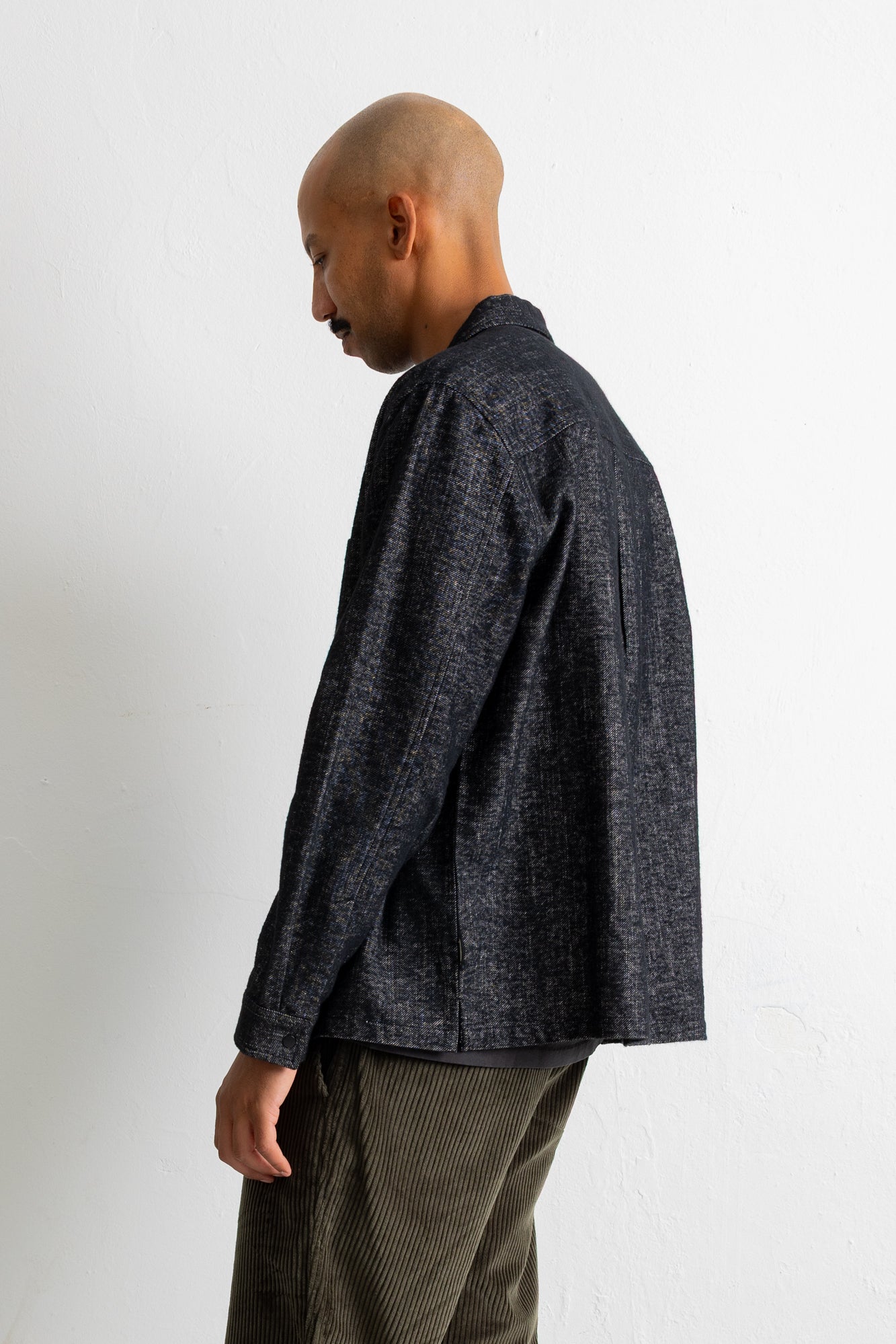 A man shown from the side profile wearing a dark charcoal gray textured long-sleeve button-up shirt and olive green corduroy trousers, standing against a plain off-white wall.