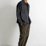 A man wearing a dark gray textured overshirt, olive green corduroy pants, and black canvas sneakers, standing against a white wall.