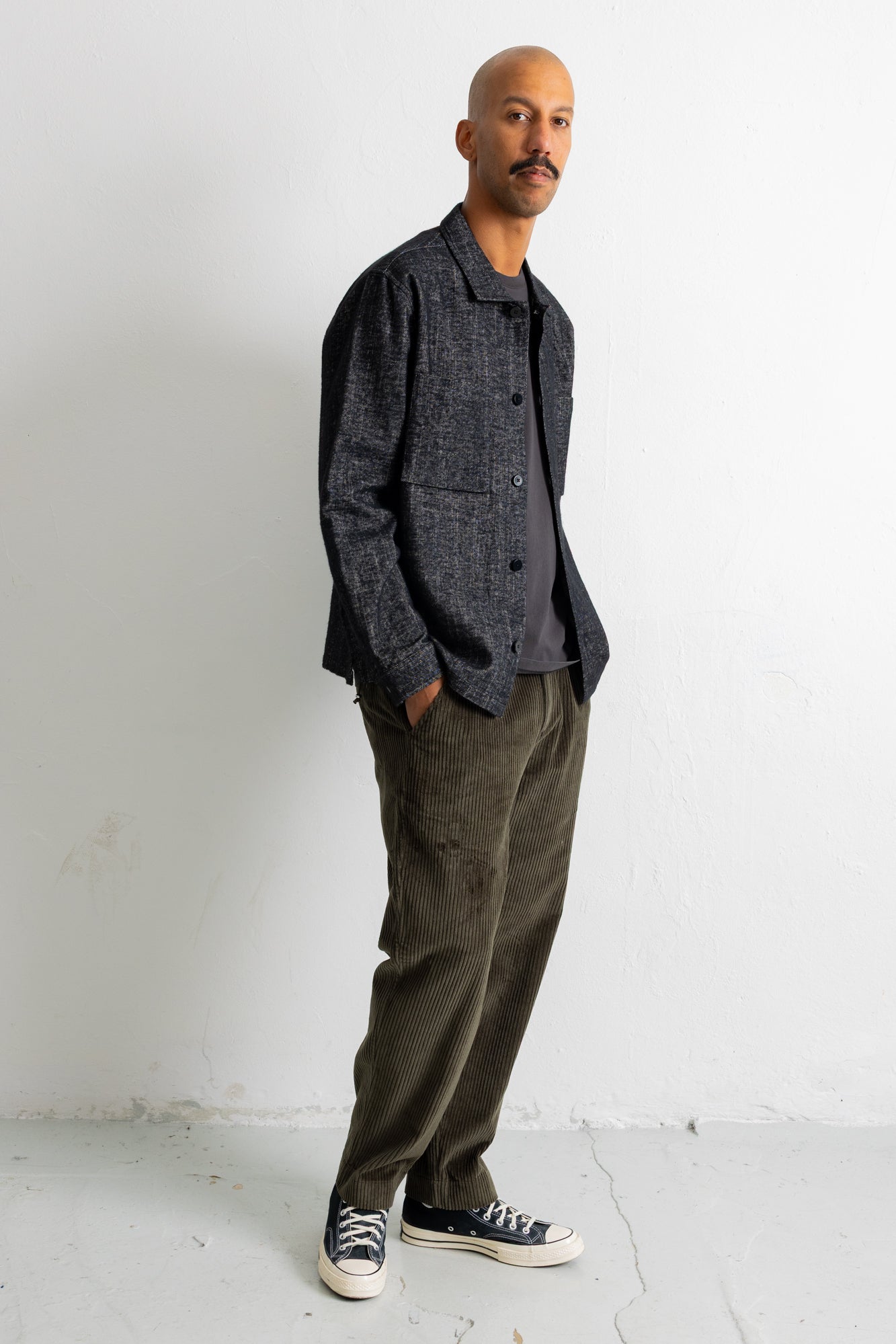 A man wearing a dark gray textured overshirt, olive green corduroy pants, and black canvas sneakers, standing against a white wall.