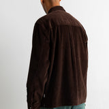 A man viewed from behind wearing a dark brown corduroy shirt jacket and teal pants, standing against a plain white wall.