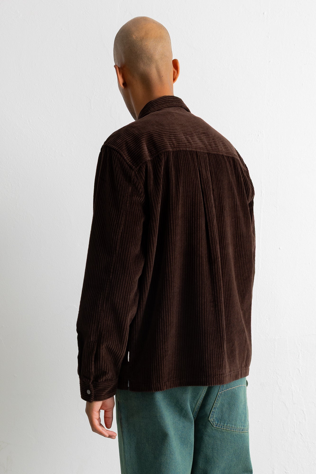 A man viewed from behind wearing a dark brown corduroy shirt jacket and teal pants, standing against a plain white wall.