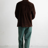 A bald man viewed from behind wearing a dark brown corduroy jacket, green trousers, and black-and-white sneakers, standing against a plain white wall.