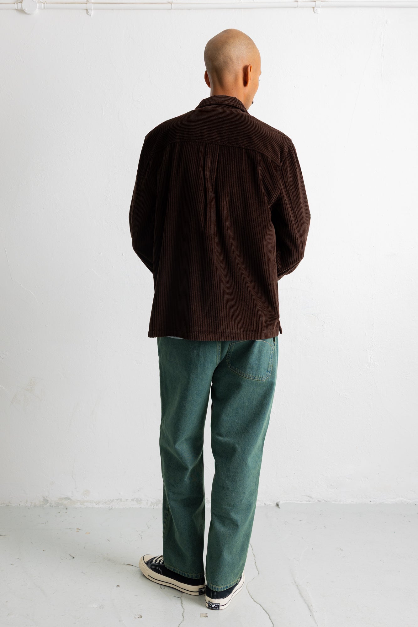 A bald man viewed from behind wearing a dark brown corduroy jacket, green trousers, and black-and-white sneakers, standing against a plain white wall.