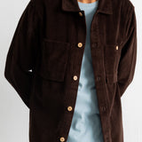 A man wearing a dark brown corduroy button-up shirt jacket over a light blue t-shirt and blue jeans, shown from the neck down against a white wall.