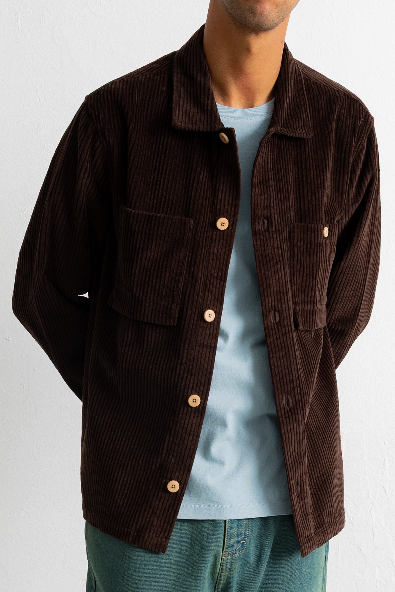 A man wearing a dark brown corduroy button-up shirt jacket over a light blue t-shirt and blue jeans, shown from the neck down against a white wall.