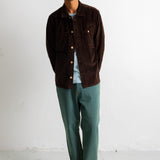 A man stands against a white wall wearing a dark brown corduroy jacket over a light blue t-shirt, paired with green trousers and black-and-white sneakers.