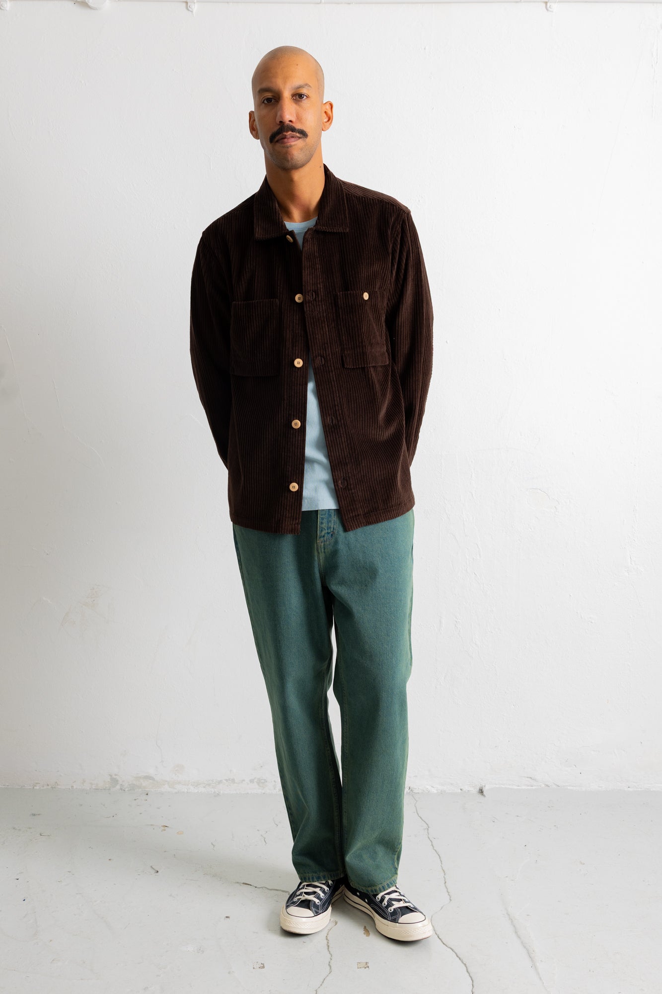 A man stands against a white wall wearing a dark brown corduroy jacket over a light blue t-shirt, paired with green trousers and black-and-white sneakers.