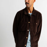 A man wearing a dark brown corduroy button-up shirt jacket over a light blue t-shirt and teal jeans, standing against a plain white wall.