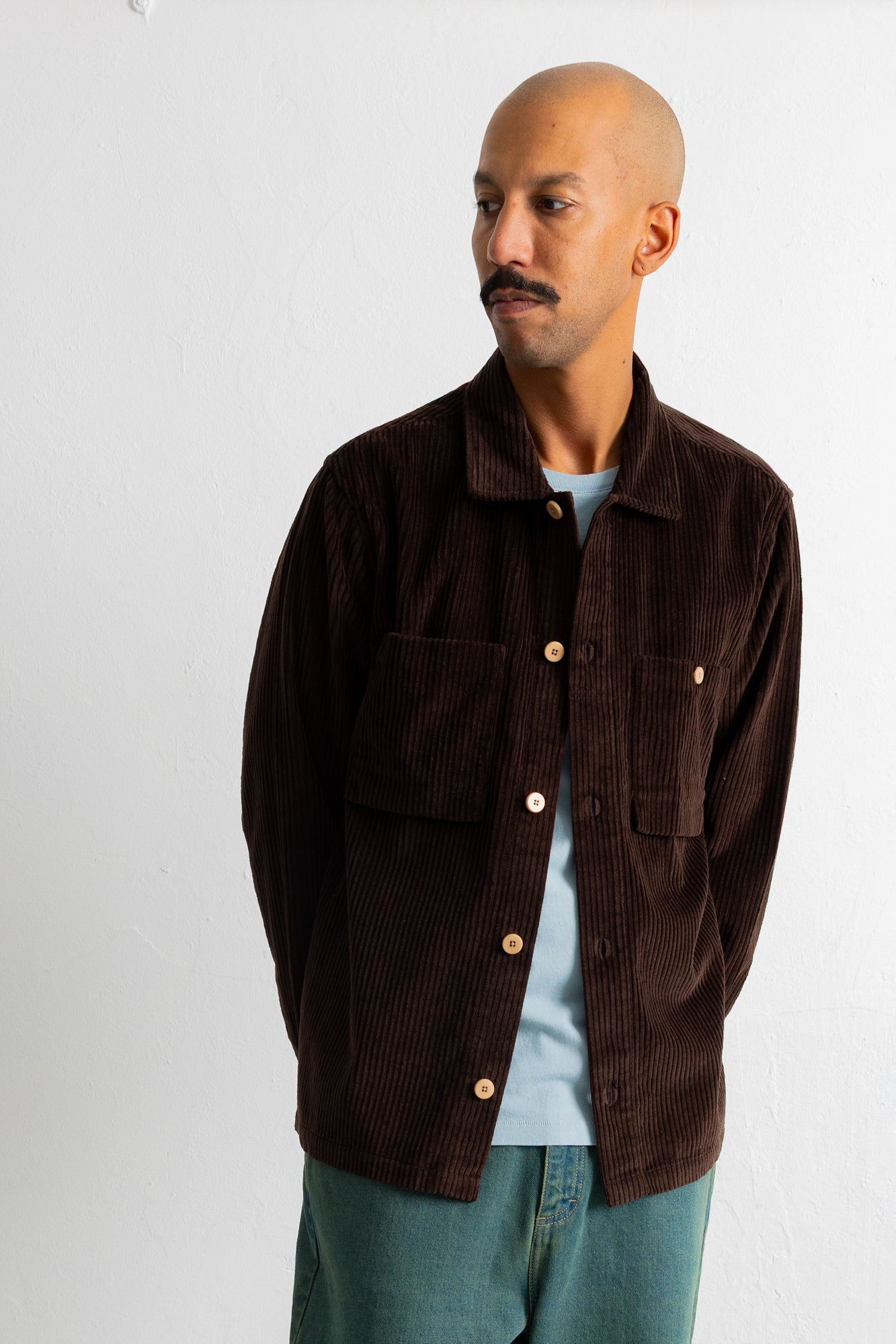A man wearing a dark brown corduroy button-up shirt jacket over a light blue t-shirt and teal jeans, standing against a plain white wall.