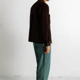 A man stands in profile wearing a dark brown corduroy jacket, teal green jeans, and navy canvas sneakers against a plain white studio wall.