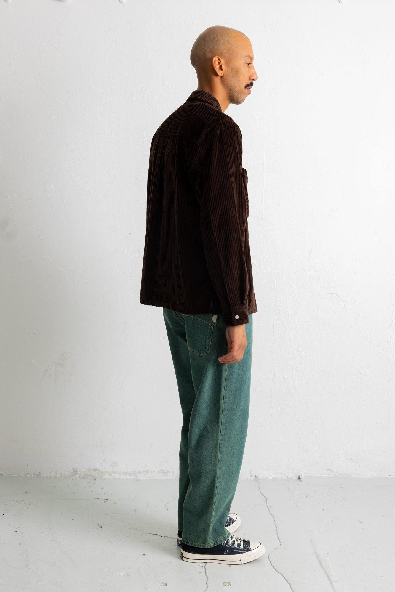 A man stands in profile wearing a dark brown corduroy jacket, teal green jeans, and navy canvas sneakers against a plain white studio wall.