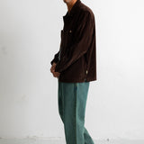 A man wearing a dark brown corduroy jacket, teal wide-leg jeans, and black canvas sneakers, standing in profile against a white studio wall.