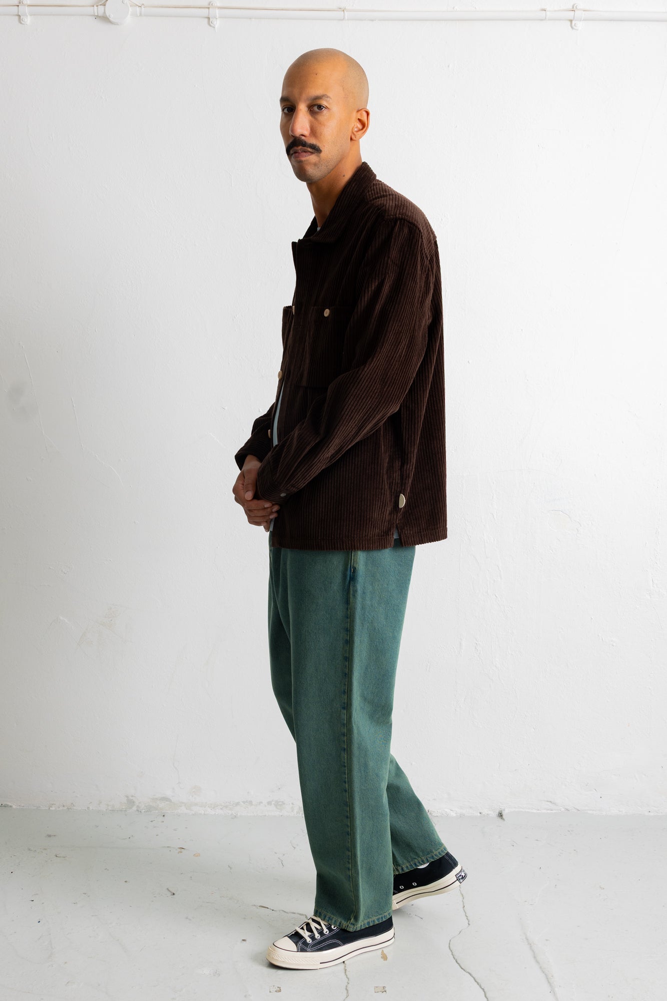A man wearing a dark brown corduroy jacket, teal wide-leg jeans, and black canvas sneakers, standing in profile against a white studio wall.