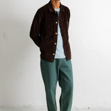 A man wearing a dark brown corduroy button-up jacket over a light blue t-shirt, paired with green trousers and black canvas sneakers, standing against a white wall.