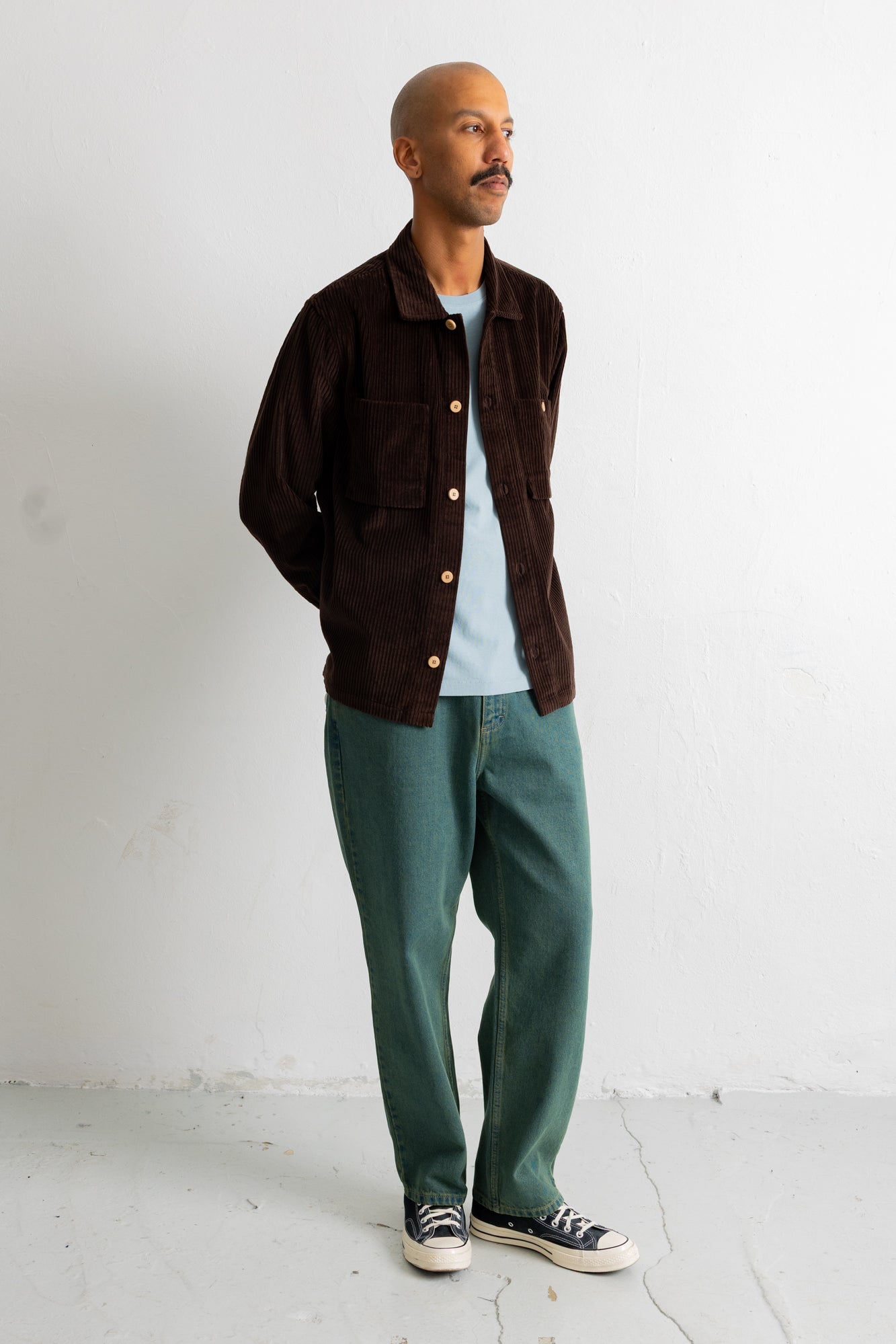 A man wearing a dark brown corduroy button-up jacket over a light blue t-shirt, paired with green trousers and black canvas sneakers, standing against a white wall.