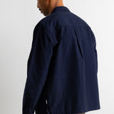A man shown from the back wearing a dark navy blue overshirt or jacket with pleated back detailing, paired with brown trousers, against a plain white wall.