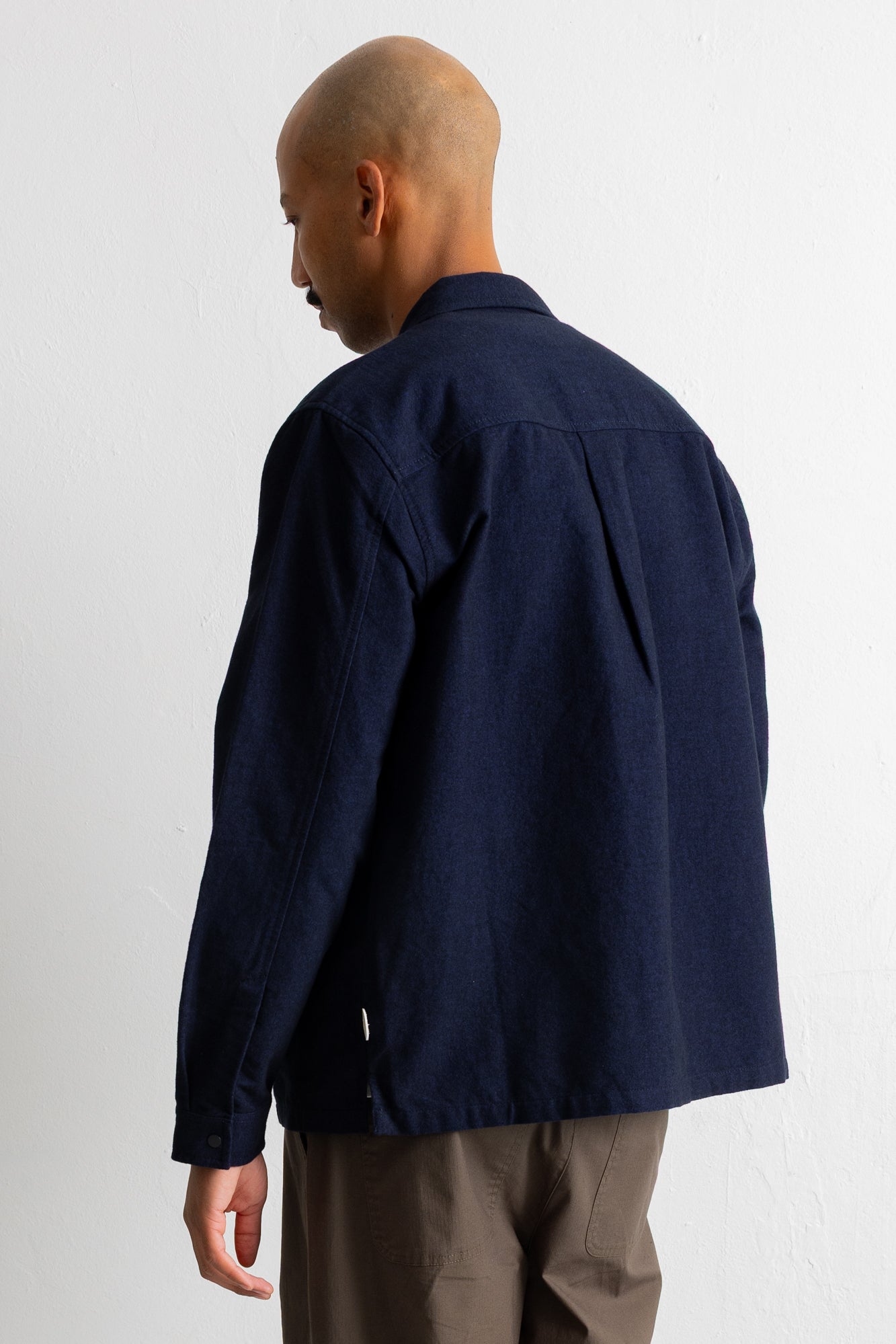 A man shown from the back wearing a dark navy blue overshirt or jacket with pleated back detailing, paired with brown trousers, against a plain white wall.