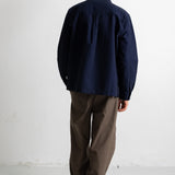 A man viewed from behind wearing a dark navy blue long-sleeve button-up shirt and brown trousers, standing against a plain white wall.